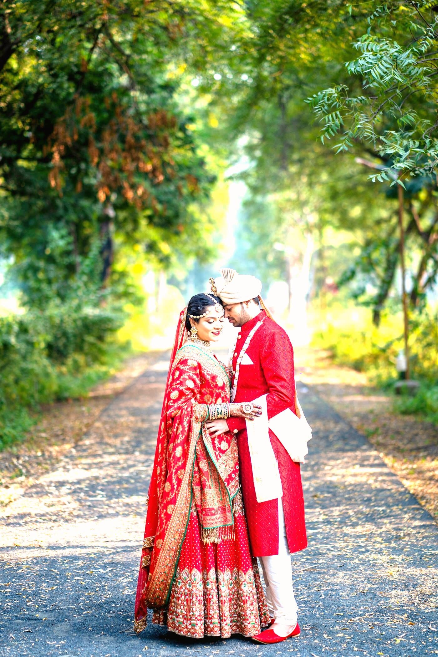 A bride and groom in red standing together on a tree-lined path in dappled sunlight