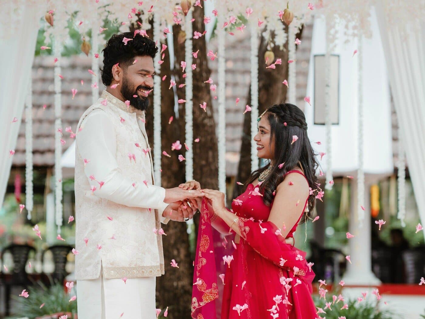 A couple in cream and red facing each other under falling flower petals in daylight