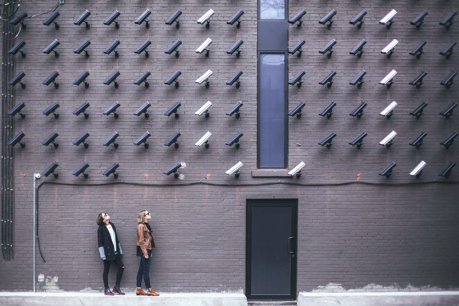 Two figures dwarfed by a facade covered in surveillance cameras